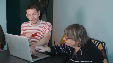 Yetişkin oğlu, yaşlı annesine laptop ve kredi kartıyla online ödeme yapmayı öğretiyor. Dijital bankacılık, finansal teknoloji öğrenimi. Nesiller arası eğitim ve aile desteği. Çevrimiçi