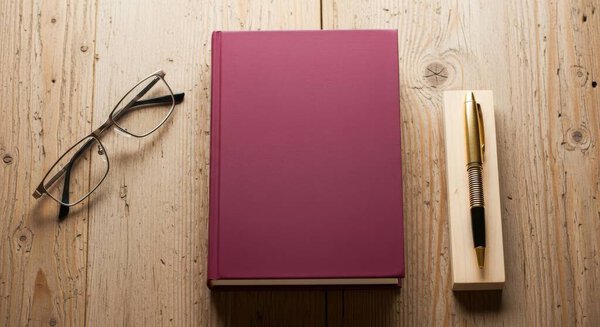 A burgundy book is placed on a wooden table, accompanied by a pen and glasses. The scene is captured from a top view, presented in a flat lay style. Perfect for education.
