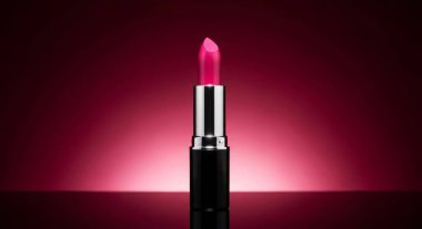 Canlı pembe bir ruj tüpü, yumuşak ışıkla aydınlatılmış koyu kırmızı bir arka fonda göze çarpar. Bu görüntü güzelliğin ve kozmetik zarafetin özünü yakalar..