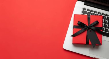 Canlı kırmızı bir hediye kutusu, parlak siyah bir kurdele ile süslenmiş, bir dizüstü bilgisayarın klavyesinde, aynı kırmızı arkaplana karşı durarak görsel olarak çarpıcı bir kompozisyon oluşturuyor..
