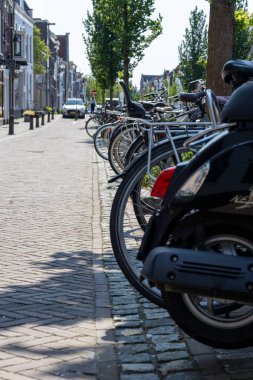 Bir sıra motosiklet ve bisiklet yan yana dizilmiş, kaldırıma park edilmiş, tuğla bir yolun yanında.