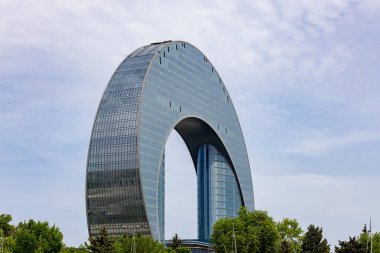 Dairesel şekilli ve bir sürü cam pencereli büyük bir bina. Bina ağaçlarla çevrili ve arka planda bulutlu bir gökyüzü var.