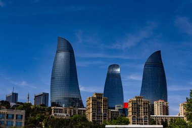 Arka planda kırmızı çatılı üç uzun bina. Binalar çok yüksek ve eşsiz bir tasarıma sahipler. Gökyüzü açık ve mavidir ve gökyüzünde hiç bulut yoktur.