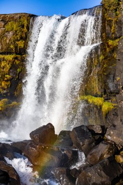 Sisin içinde gökkuşağı olan bir şelale. Su berrak ve kayalar gri. Şelale yeşil yosunlarla çevrili ve gökyüzü mavi.