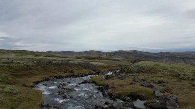 İzlanda manzarası. Üstü bulutlu bir gökyüzüyle çimenli bir tarladan akan bir nehir. Sahne sakin ve huzurlu, su yavaşça akıyor ve otlak sakin bir zemin sağlıyor.