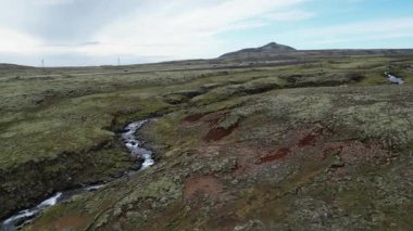 İzlanda manzarası. Bir nehir, üzerinde açık mavi gökyüzü olan çimenli bir ovanın içinden akar. Sahne huzurlu ve sakin, su peyzaj boyunca sakince akıyor.