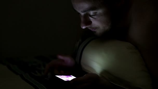 Young man using smartphone in the bed under blanket by night — Stock Video