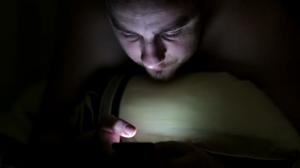 Young man using smartphone in the bed under blanket by night — Stock Video