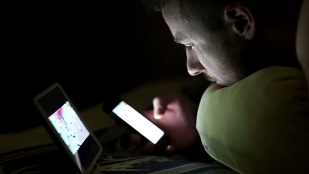 Молодой человек с планшетом и смартфоном в постели ночью — стоковое видео
