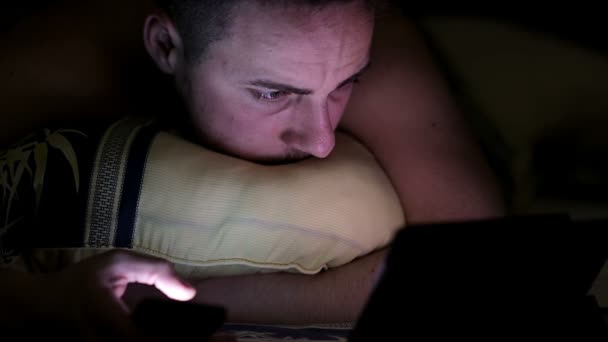 Молодой человек с планшетом и смартфоном в постели ночью — стоковое видео