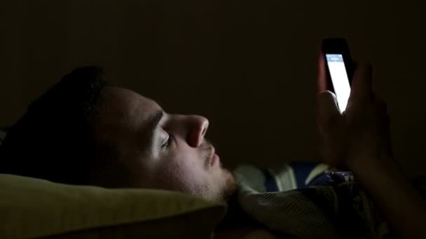 Jovem usando smartphone na cama sob cobertor à noite — Vídeo de Stock