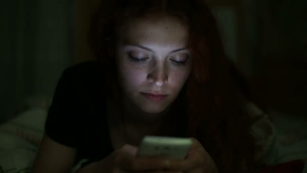 Jovem mulher usando smartphone deitado na cama em casa à noite — Vídeo de Stock