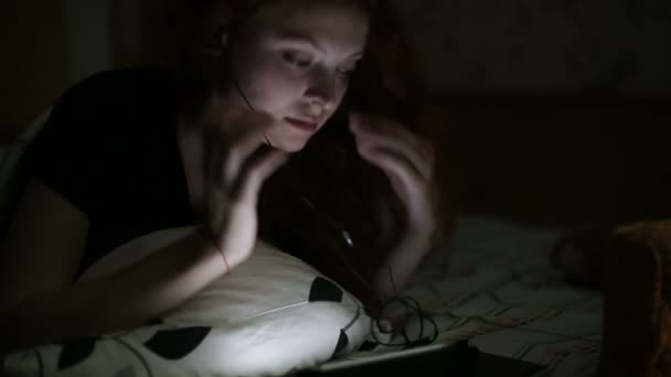 Молодая женщина слушает музыку с табличкой, лежащей на кровати в ночное время дома — стоковое видео