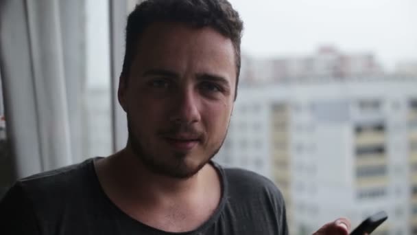 Молодой человек пьет кофе и пользуется смартфоном на балконе — стоковое видео