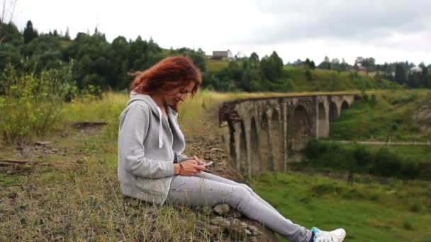 La chica escribe sms sentado cerca del viejo viaducto en las montañas — Vídeo de stock