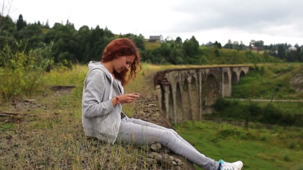 Het meisje schrijft sms zitten in de buurt van het oude viaduct in de bergen — Stockvideo