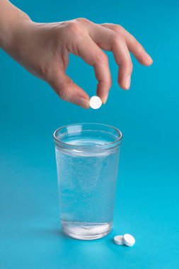 Bir bardak su, vitamin, hap ve tablet. Açık mavi dikey arka plan. Kadın eli bir hap atar..