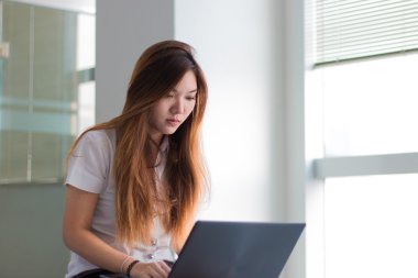 Asyalı öğrenci pencere yakın bir binada bir dizüstü bilgisayar üzerinde çalışan