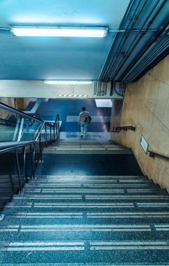 Yetişkin bir adam karanlık bir metro istasyonunda beton merdivenlerden iniyor. Altgeçitte mavi floresan lambalar ve endüstriyel borular var..