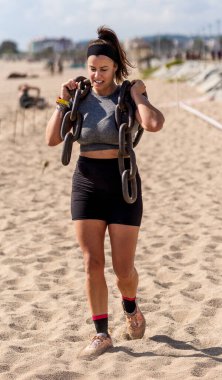 Odaklanmış bir bayan atlet güneşli bir günde kumlu sahilde omuzlarında ağır zincirler taşımaya çalışır..