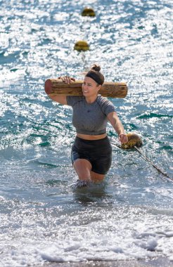 Güçlü ve kararlı genç bir bayan sporcu, engelli bir pistte köpüklü deniz suyundan ağır ahşap bir kütük taşıyor..