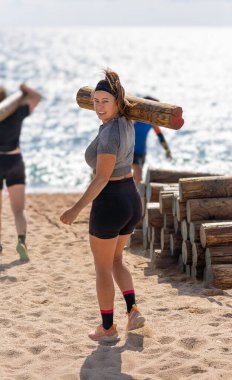 Güçlü, gülümseyen bir bayan atlet omzunda ağır bir kütük taşıyor, kameraya bakıyor, sahildeki engelli bir pistte..