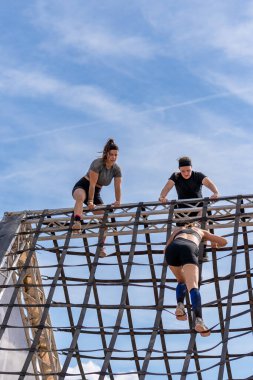 Üç bayan sporcu dik, piramit şeklinde bir ağa tırmanıyor mavi gökyüzünün arka planına karşı..