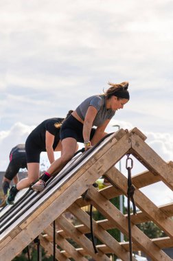 Kararlı bir bayan sporcu ve diğer yarışmacılar, açık hava engelli yarışı sırasında zirvedeki tahta A-çerçeve engelini aşıyorlar..