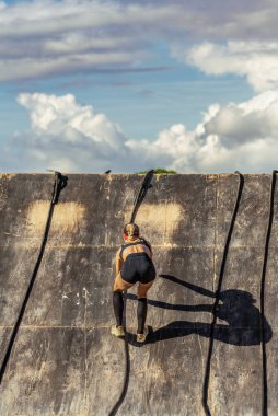 Güçlü bir bayan sporcunun, bulutlu mavi gökyüzünün altındaki dayanıklılık yarışı sırasında ahşap bir engel duvarına tırmanmasının görüntüsü..