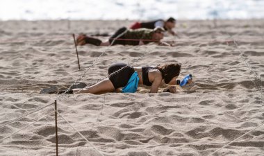 Genç, esmer tenli bir sporcu arka planda diğer sporcularla birlikte açık hava engelli parkurunda dikenli tellerin altında kumsalda emeklemeye çalışıyor..