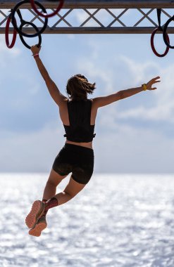 Sallanan halkalar üzerinde dengede duran genç bir atletin arkasından, engelli bir pistte başka bir ringe ulaşmak için sallanırken, arka planda parlak gökyüzü ve deniz onun siluetini çiziyor..