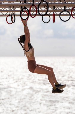 İnce kadın sporcu, deniz kenarındaki bir engelli parkurunda parıldayan okyanus sularının üzerindeki halka sahasında asılı duruyor..