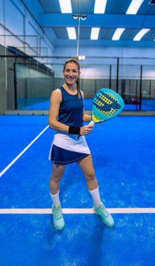 Padel bir oyuncu padel etek giyiyor ve üstte mutlu poz veriyor, gülümsüyor, mavi bir kortta raketle duruyor..