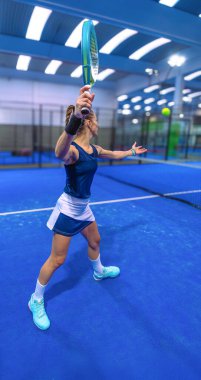 Dişi Padel oyuncusu raketini kaldırıyor. Topun yaklaşmasını bekliyor. Mavi kortta topu yere sermek için..