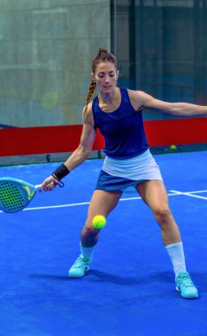 Kararlı bir bayan Padel oyuncusu topun ona yaklaşmasını izliyor ve mavi bir iç sahada düşük bir atış yapıyor..