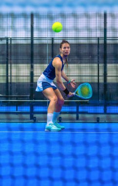 Padel maçına odaklanmış genç bir kadının Padel kortunun ağıyla, kapalı sahada kendisine doğru yaklaşan topun ters vuruşunu yapmaya hazırlanıyor..