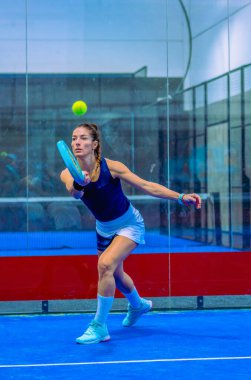 Kararlı genç bir Padel oyuncusu, kapalı bir mavi kortta bir padel maçında son saniye vuruşunu gerçekleştiriyor..