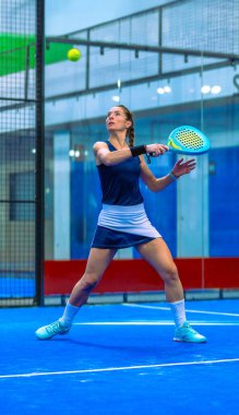 Atletik bayan Padel oyuncusu, mavi kapalı bir padel kortunda topun yüksekliğinde görsel takibiyle teknik bir ters vuruş yapıyor..