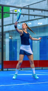 Atletik padel oyuncusu, kapalı sahada oynanan yoğun bir Padel maçı sırasında çok yüksek bir top atışı ve büyük bir gerilim ve teknik ile vuruşu gerçekleştiriyor..