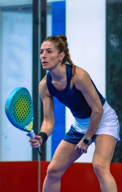 Güzel bir bayan Padel oyuncusunun orta boy çekimi. Odaklanmış ve hazır. Kapalı alanda oynanan maçta dizleri ve gözleri sabitlenmiş..