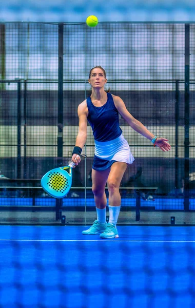 Kararlı bir bayan Padel oyuncusu mavi bir iç sahada topa vurmak için yukarı bakıyor..