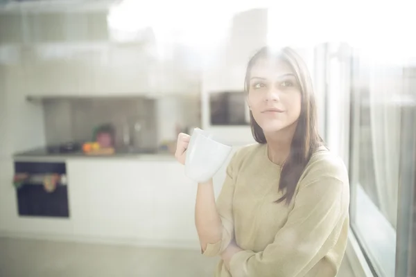 Woman enjoying,holding cup of hot beverage,coffee or tea.Enjoying her morning coffee in the kitchen.Savoring a cup of coffee in bliss and appreciation.Looking trough the window — Stock Photo, Image