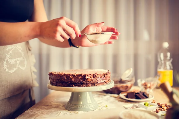 Hausfrau tragen Schürze macht den letzten Schliff auf Geburtstag Dessert Schokoladenkuchen. Frau macht hausgemachten Kuchen mit einfachem Rezept, Streuen Puderzucker auf der Oberseite — Stockfoto