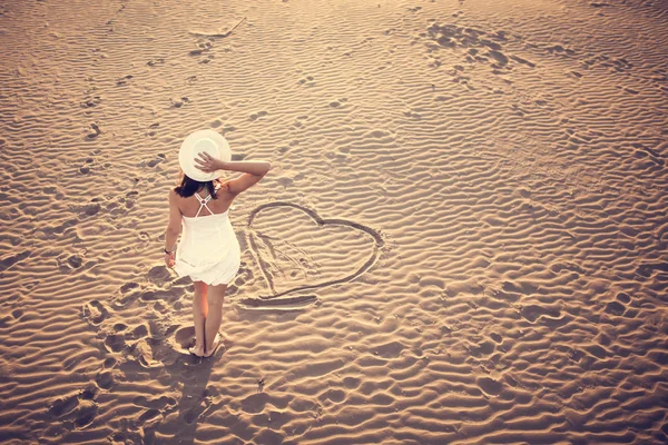 Жінка на пляжі робить серце на піску. Молода жінка ходить на піску в білій сукні. . — стокове фото