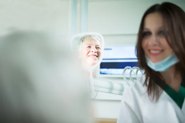 Старуха посещает дантиста, заботится о зубах. Молодая женщина-дантист показывает бабушке зубы, отражение в зеркале. Врач-дантист разговаривает со старшей женщиной. — стоковое фото