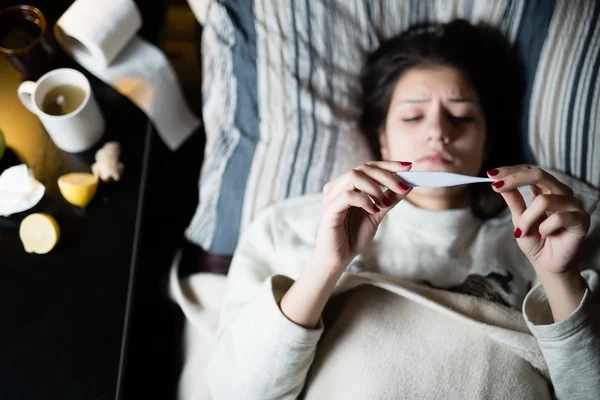 Mujer joven enferma en la cama en casa teniendo gripe, midiendo la temperatura.Termómetro para comprobar la temperatura para la fiebre.Flu.Virus.Mujer enferma acostada en la cama.Enfoque en el termómetro — Foto de Stock