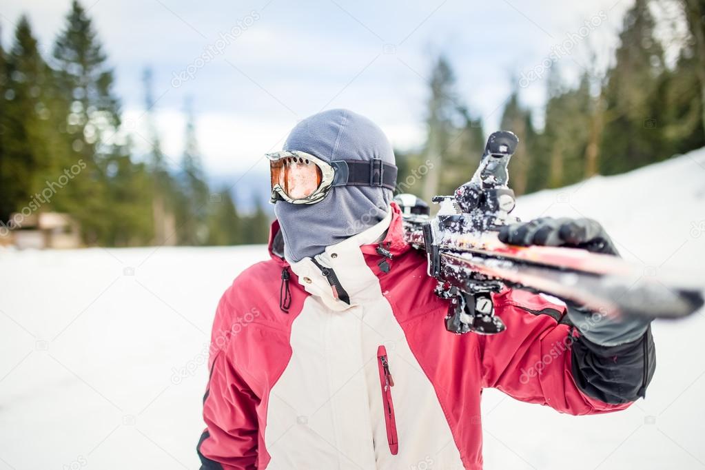 Young man holding ski at ski resort.Skier holding skis looking at the mountains.Side view of handsome skier man with mask and holding ski equipment