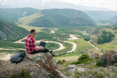 Dağın tepesindeki bilgisayarında çalışan yetişkin bir adam. Uzaktan çalışma konsepti. Gezgin yaz tatillerinde dağların tadını çıkarırken uzaktan çalışıyor..