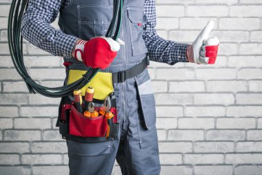 Elektrikçi üniformalı, yuvarlanmış kablolar tutan ve arka planda başparmağını gösteren.. 