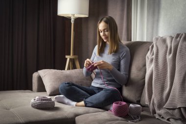 Genç ve güzel bir kadın kanepede oturuyor ve evde örgü örüyor. Kadın hobisi..